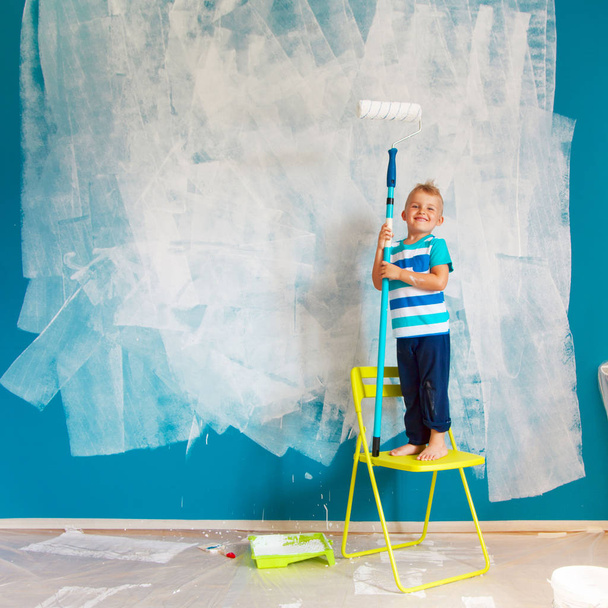 The child paints the blue wall with a roller. The boy holds a large brush for painting. Home repairs. - Foto, Imagem