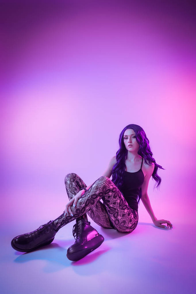 Женщина с длинными фиолетовыми волосами в черном нижнем белье, колготки, позирующая в привлекательном положении в ярких неоновых синих и розовых огнях в студии. Концепция моды
 - Фото, изображение