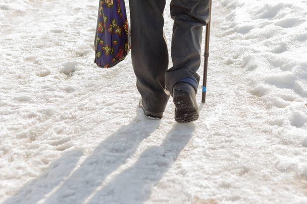 Ein älterer Mann mit Stock und Einkaufstasche geht durch den schmutzigen, lockeren Schnee - Foto, Bild