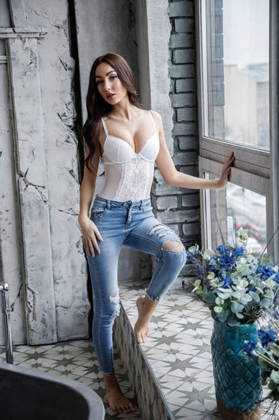 Studio portrait of beautiful brunette woman wearing white lingerie and blue jeans. Charming beauty model posing at apartments interior   - Фото, изображение