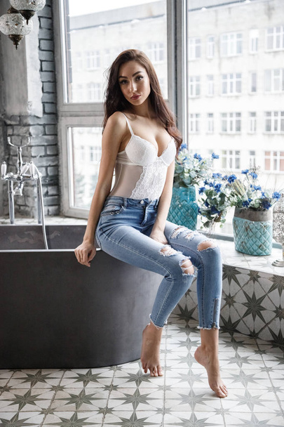 Studioporträt der schönen brünetten Frau in weißen Dessous und blauen Jeans. Charmante Schönheit Modell posiert in Wohnungen Interieur   - Foto, Bild