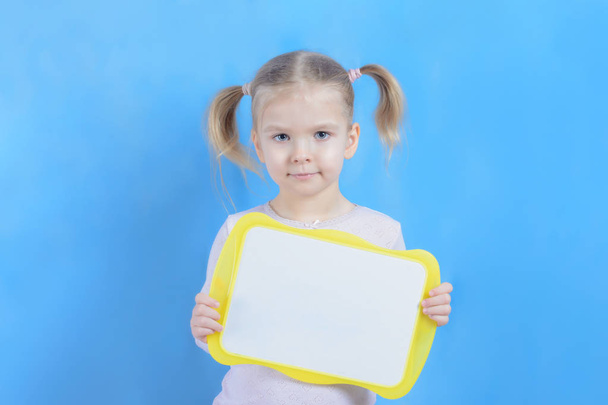 Glimlachend klein meisje met blond haar dat leeg teken op blauwe achtergrond houdt - Foto, afbeelding
