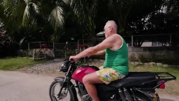 老人は、夏休みの間の田舎でオートバイを運転します。旅行をしながら熱帯村の通りにバイクに乗ってサングラスで年配の男性. - 映像、動画