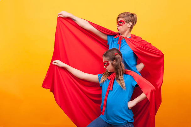 Young mother and her little girl dressed like heroins holding their red cape in the air over yellow background. - Фото, изображение