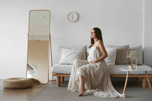 Beautiful brown-haired pregnant model girl with blue eyes and with a pretty smile in a stylish beige dress sits on the sofa and posing at minimalist interior - Фото, изображение