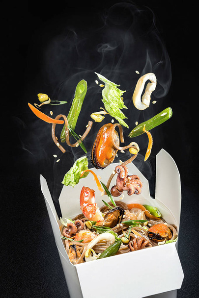 Sea food with vegetables in a box. Ingredients such as octopus, mussels, shrimp peppers, lettuce, asparagus, rice and sesame seeds are falling. Delivery of Asian food. - Photo, Image