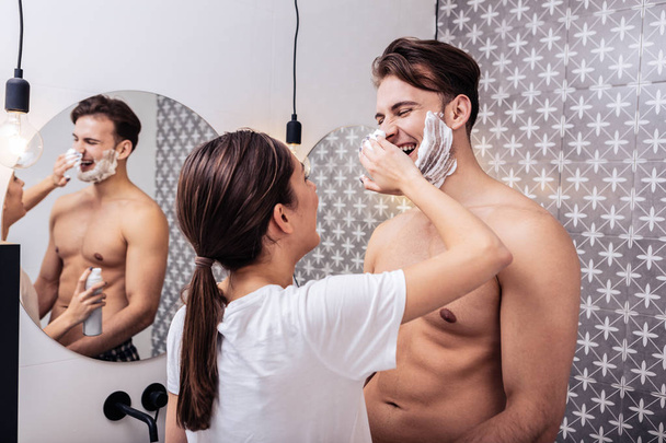 Wife putting shaving foam on his nose standing in the bathroom - Fotó, kép
