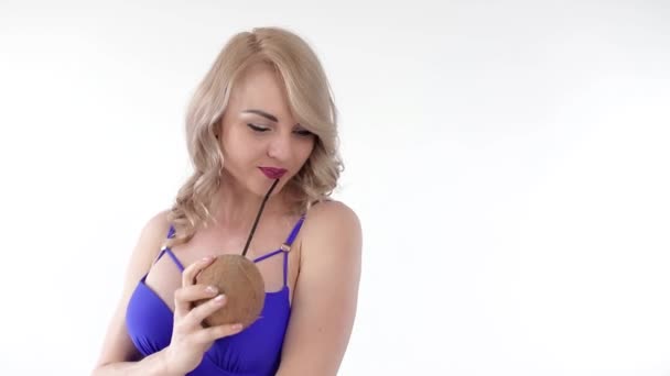 Portrait of a young attractive blonde woman wearing a blue beachwear drinking juice from a coconut through a straw, wind is blowing in her face. Looking at the camera, isolated on white background. - Footage, Video