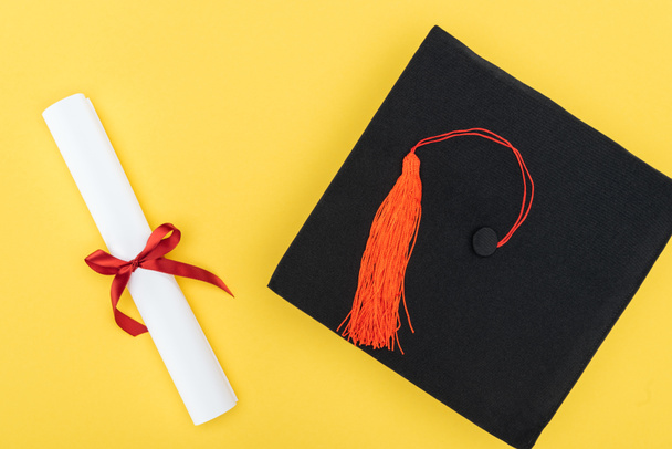 Top view of academic cap and diploma with red ribbon isolated on yellow - Photo, Image