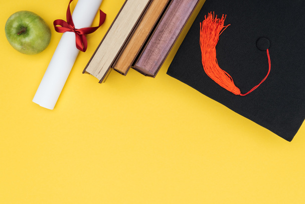 Pohled shora knih, jablko, akademické čepice a diplom na žlutém povrchu - Fotografie, Obrázek