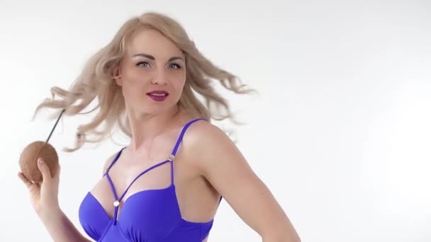 Portrait of a young sensual blonde woman dressed in blue swimwear drinking juice from a coconut through a straw, wind is blowing in her face. Looking at the camera, isolated on white background. - Záběry, video