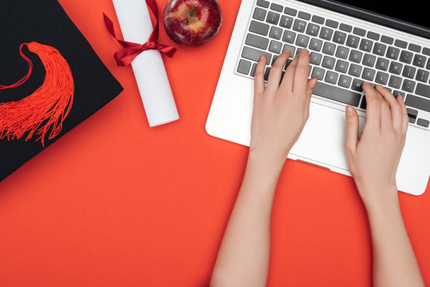 Vista recortada de chica con gorra académica, diploma y manzana utilizando el ordenador portátil en la superficie roja
 - Foto, Imagen
