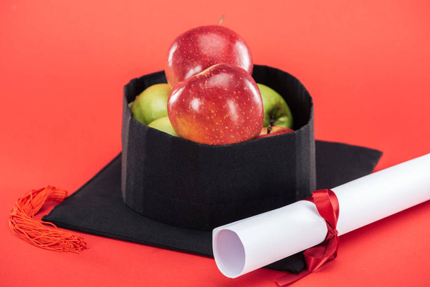 Ακαδημαϊκό καπάκι με μήλα και δίπλωμα σε κόκκινο επιφάνεια - Φωτογραφία, εικόνα