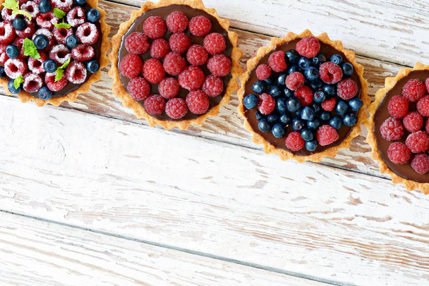 Raspberry and blueberry tartlets with chocolate ganache, fresh berries and mint leaves, selective focus. Fresh fruit tart on white background, freshly homemade fruit cake on a table - Foto, imagen