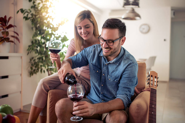 Νεαρό ζευγάρι αγαπώντας, πίνοντας κόκκινο κρασί στο σαλόνι τους. - Φωτογραφία, εικόνα