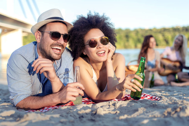 Joven hermosa pareja divirtiéndose en la playa, bebiendo cerveza y sonriendo
. - Foto, imagen