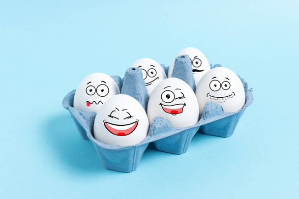 Easter eggs with funny face in carton tray on blue background. Minimal concept. - Фото, изображение