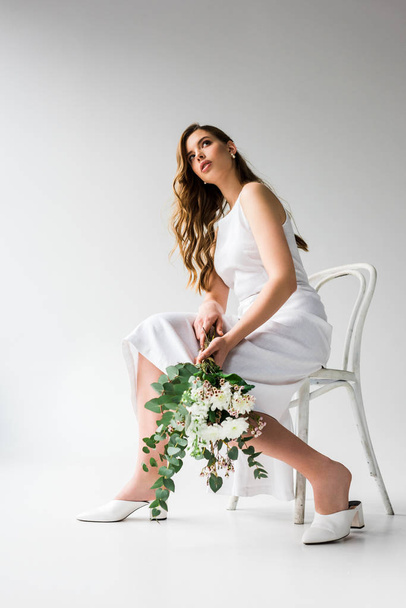 vista de ángulo bajo de la mujer en vestido sentado en la silla y la celebración de ramo de flores con hojas de eucalipto en blanco
  - Foto, Imagen