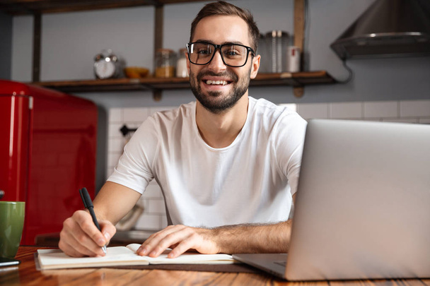 Photo of smiling man writing down notes while using silver lapto - Foto, Imagem
