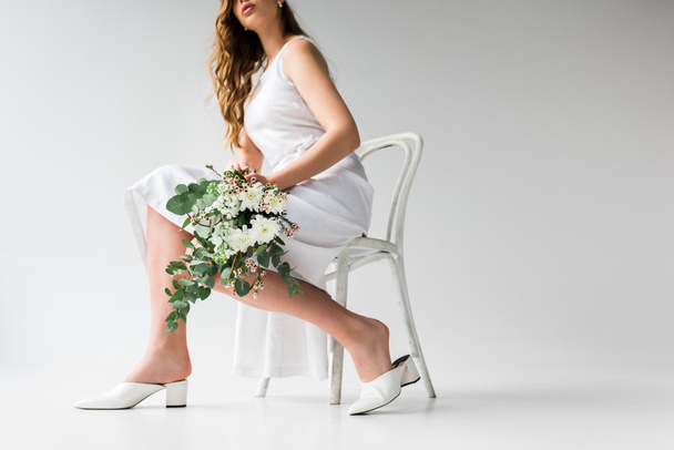 cropped view of woman in dress sitting on chair and holding bouquet of flowers with eucalyptus leaves on white  - Photo, Image