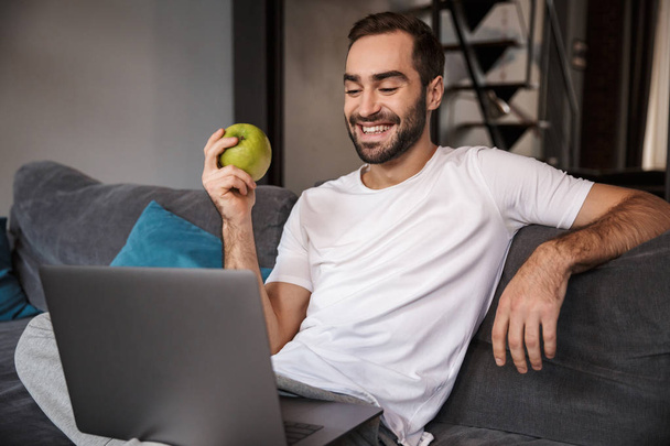 Photo of happy bachelor using silver laptop while sitting on cou - Foto, Bild