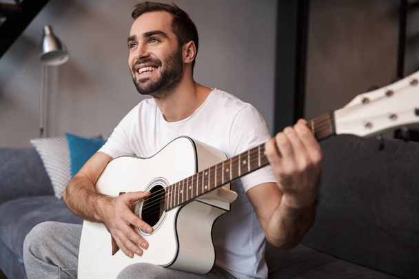 Photo of young man playing acoustic guitar while sitting on sofa - Foto, Bild