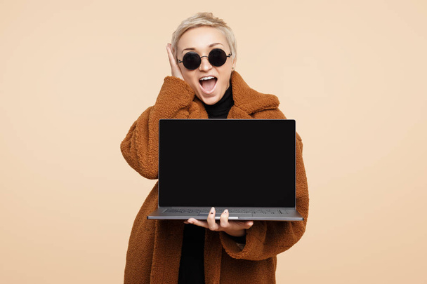 Shocked young hipster woman with blonde short hair wearing a coat and sunglasses holding a computer or laptop isolated over beige background. - Foto, Imagem