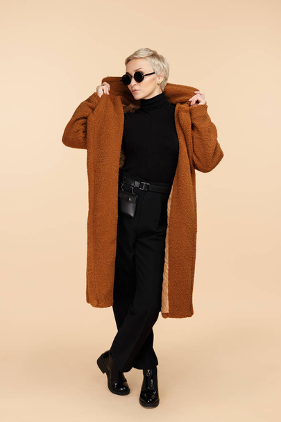 Charming young hipster woman with blonde short hair wearing a coat and sunglasses posing isolated over beige background. Full lengh photo. - Foto, immagini