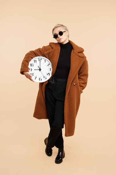 ernsthafte junge Hipsterfrau mit blonden kurzen Haaren, Mantel und Sonnenbrille, die eine Uhr vor beigem Hintergrund hält. - Foto, Bild