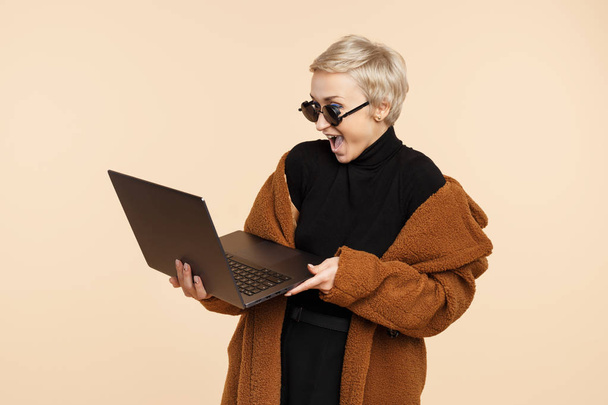 Shocked young hipster woman with blonde short hair wearing a coat and sunglasses holding a computer or laptop isolated over beige background. - Φωτογραφία, εικόνα