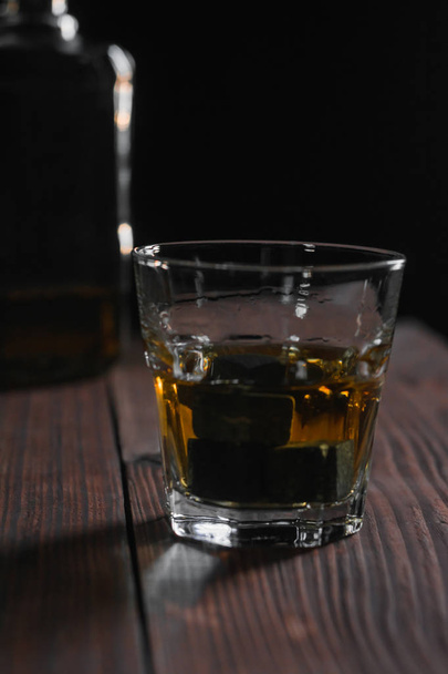 Photo contrastée verticale d'un verre de whisky et de pierres debout au premier plan sur une table en bois. En arrière-plan se trouve un fragment de bouteille
. - Photo, image