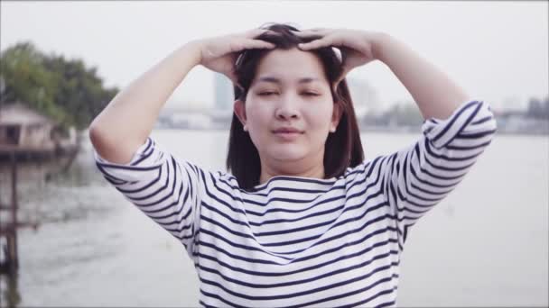Ritratto ravvicinato della giovane asiatica che sorride e usa la mano per massaggiare la testa con i capelli neri che soffiano nel vento guardando il tramonto vicino al fiume. La donna che indossa è camicia a quadri. Rallentatore
. - Filmati, video
