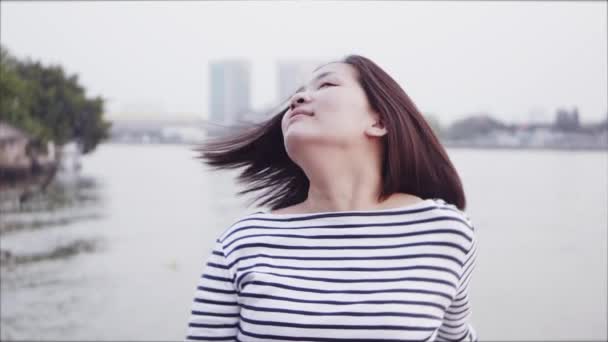 Lähikuva muotokuva nuori aasialainen nainen hymyilee ja näpäyttää päätä ja hiuksia musta hiukset puhaltavat tuulessa katselee auringonlaskua lähellä jokea. Nainen yllään on ruudullinen paita Slow Motion
. - Materiaali, video