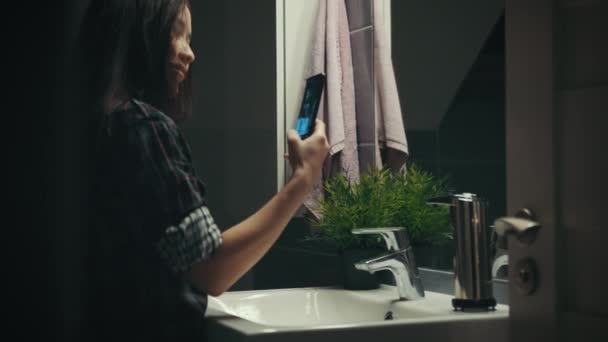 Young woman taking selfie in front of mirror in the bathroom at night, camera on slider - Felvétel, videó