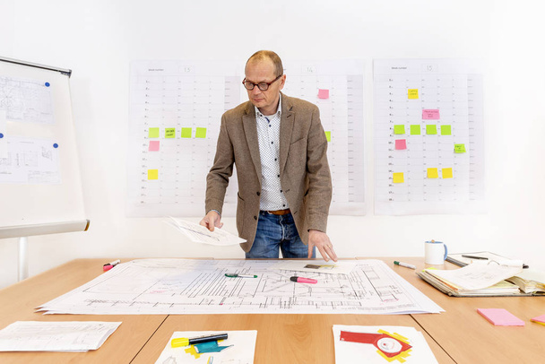 A senior managing contractor behind a desk with piles of technical drawings and sketches. On the wall behind him there is a planning board and a white board - Fotoğraf, Görsel
