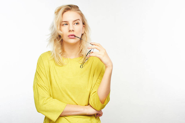 Pensive European stylish young woman in yellow clothes holding glasses near mouth, doubts what to choose, thinks, decides what to buy on white background - Фото, зображення