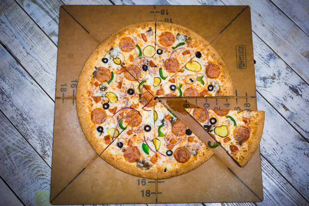 Appetizing large pizza cut into pieces, lying on the Board with numbers indicating its diameter 16cm. A piece of fresh pizza on a brown Board, hand takes pizza, cuts with a knife. - Photo, image