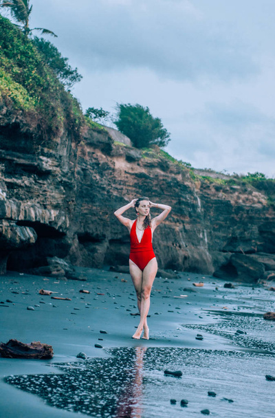 Young woman walking along the black sand beach in Bali wearing red swimsuit. - Foto, afbeelding
