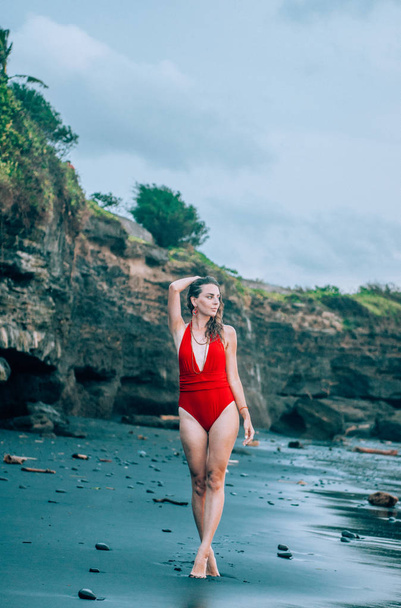 Young woman walking along the black sand beach in Bali wearing red swimsuit. - Photo, image
