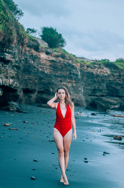 Young woman walking along the black sand beach in Bali wearing red swimsuit. - Foto, imagen
