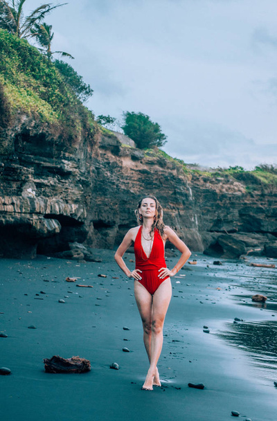 Young woman walking along the black sand beach in Bali wearing red swimsuit. - Foto, Imagen