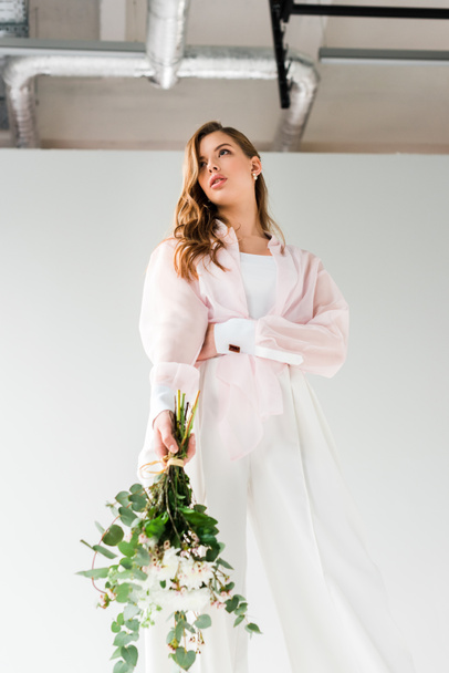 low angle view of attractive woman holding flowers with green eucalyptus leaves while standing on white  - Foto, afbeelding