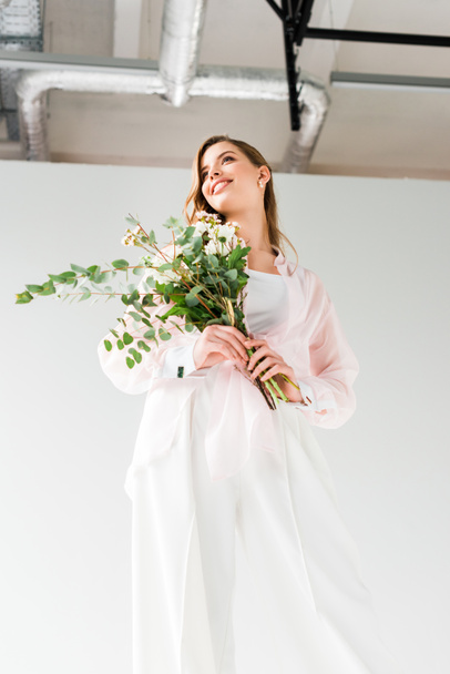 low angle view of happy woman holding flowers with green eucalyptus leaves while standing on white  - Photo, Image