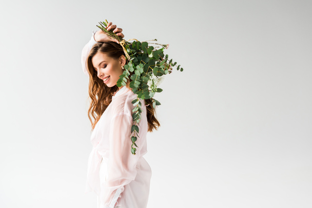 cheerful beautiful girl holding flowers with green eucalyptus leaves behind back on white  - Valokuva, kuva