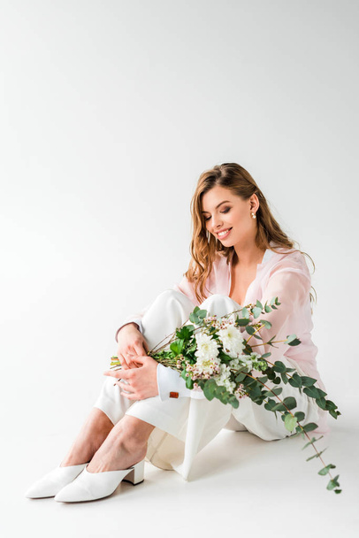 smiling young woman sitting with flowers and green eucalyptus leaves on white  - Foto, imagen