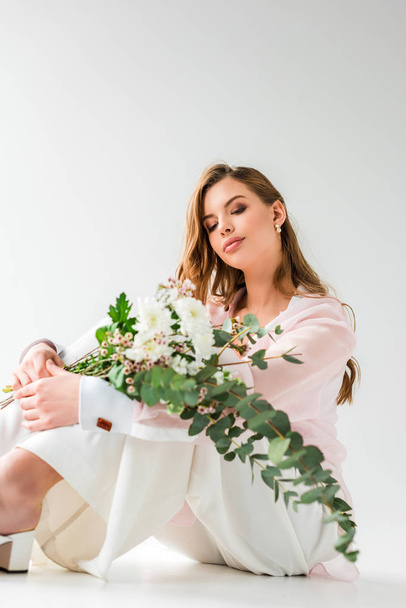 young woman looking at bouquet of flowers and green eucalyptus leaves while sitting on white  - Фото, зображення