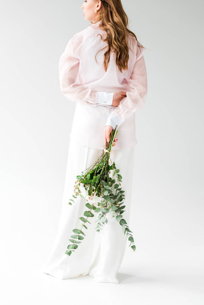 cropped view of young woman holding bouquet of flowers with green eucalyptus leaves behind back on white  - Фото, изображение