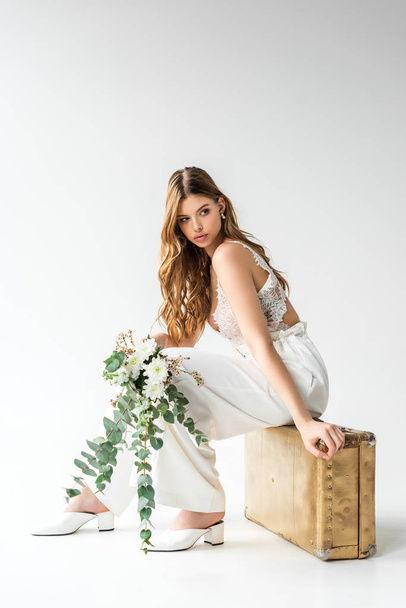 attractive girl sitting on travel bag and holding bouquet with flowers and eucalyptus leaves on white  - Valokuva, kuva