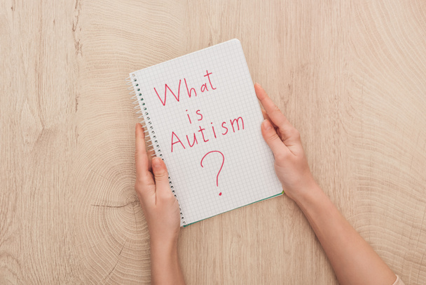 Ausgeschnittene Ansicht einer Frau, die ein Notizbuch mit der Frage nach Autismus auf einem Holztisch hält - Foto, Bild