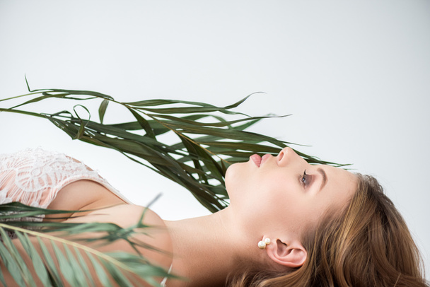 side view of attractive young woman lying near tropical palm leaves on white  - Φωτογραφία, εικόνα